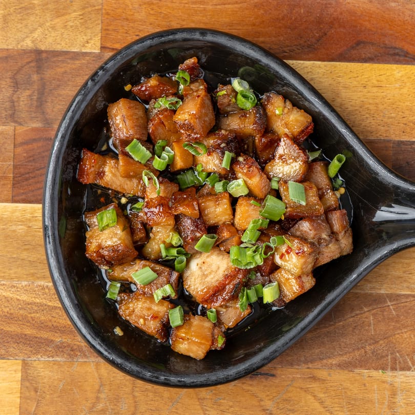 Close-up of glazed pork belly pieces topped with chopped green onions in a black dish.