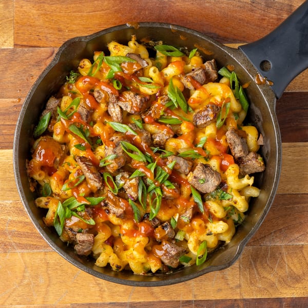 Skillet of macaroni and cheese with steak, hot sauce, and green onions on a wooden surface.