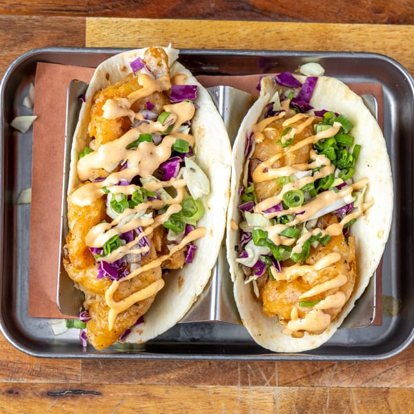Two crispy fish tacos with red cabbage, green onions, and a creamy sauce on a metal tray.