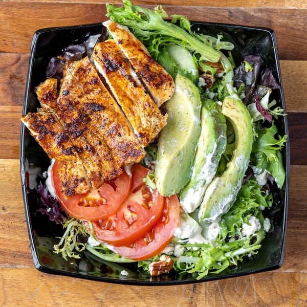 Grilled chicken salad with avocado, tomatoes, mixed greens, and dressing in a black bowl.