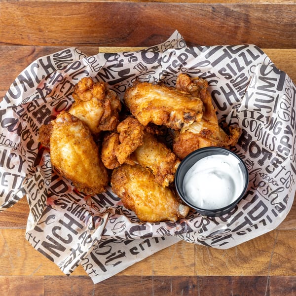 Crispy fried chicken wings and a side of ranch dressing in a paper-lined basket on a wooden table.