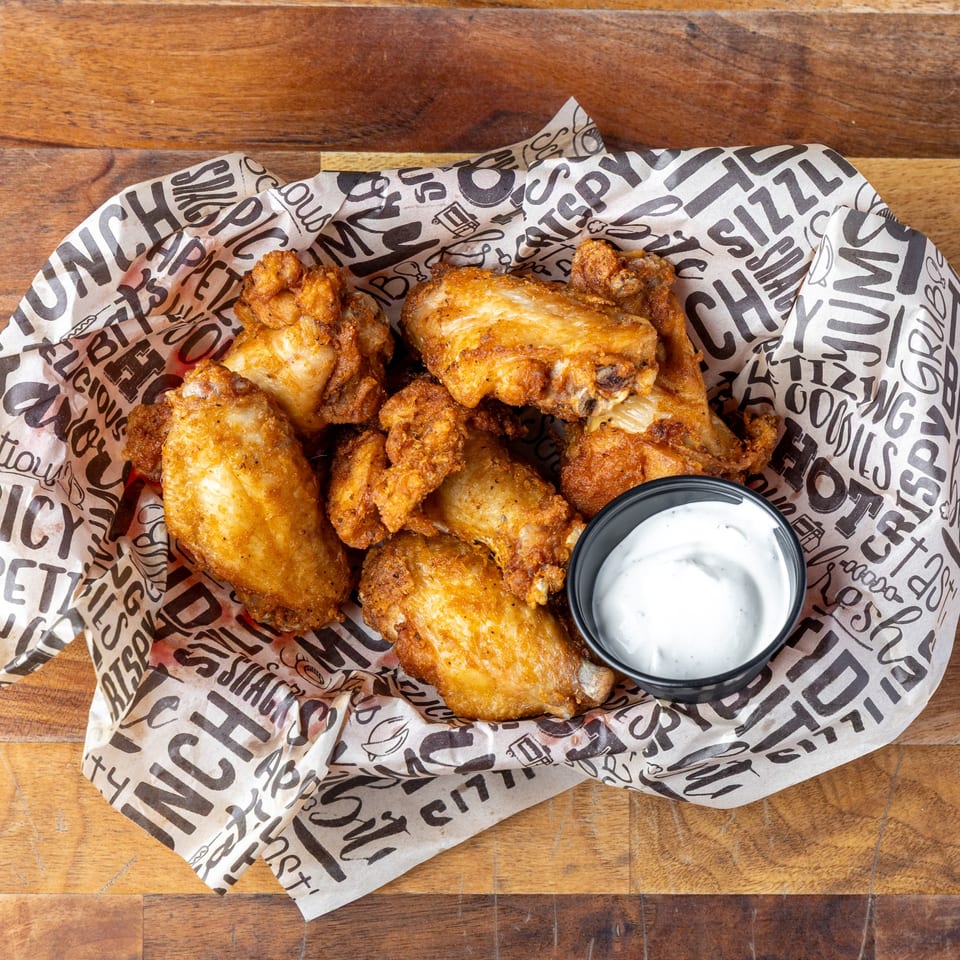 Crispy fried chicken wings and a side of ranch dressing in a paper-lined basket on a wooden table.