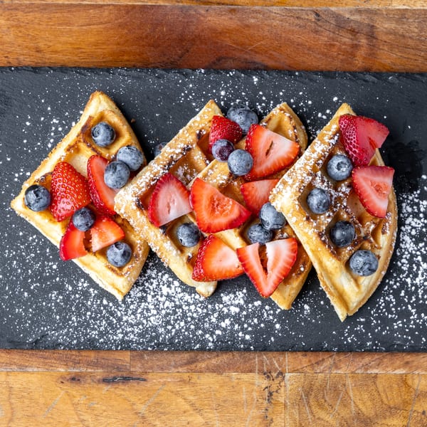 Three golden waffles topped with fresh strawberries, blueberries, and powdered sugar on a dark slate.